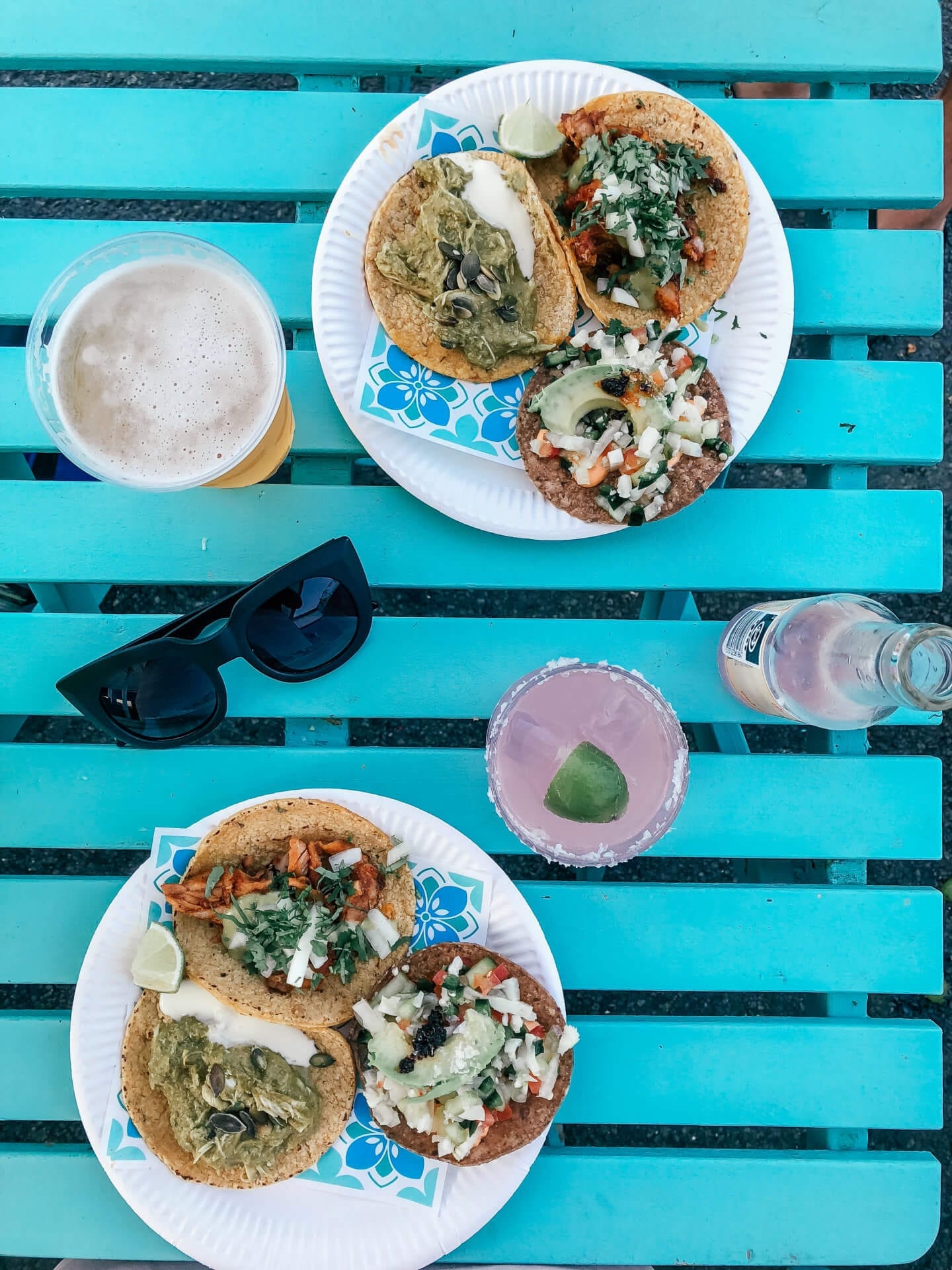 Looking down at a turquoise table with tacos, sunglasses, beer and a pink margarita