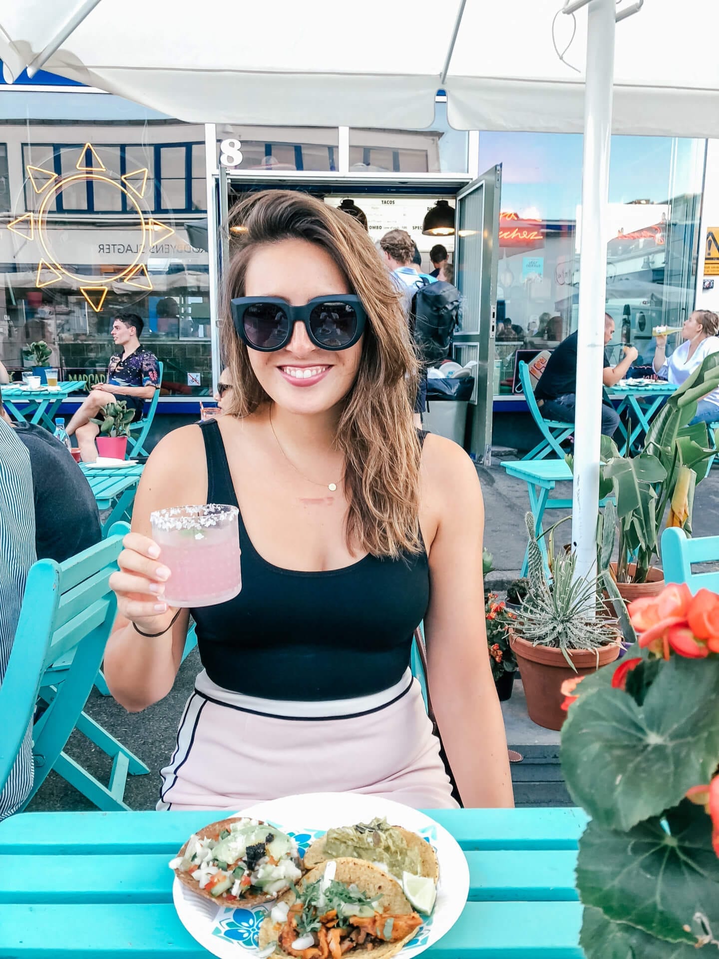 A girl with sunglasses on holding a pink margarita with a plate of tacos in front of her
