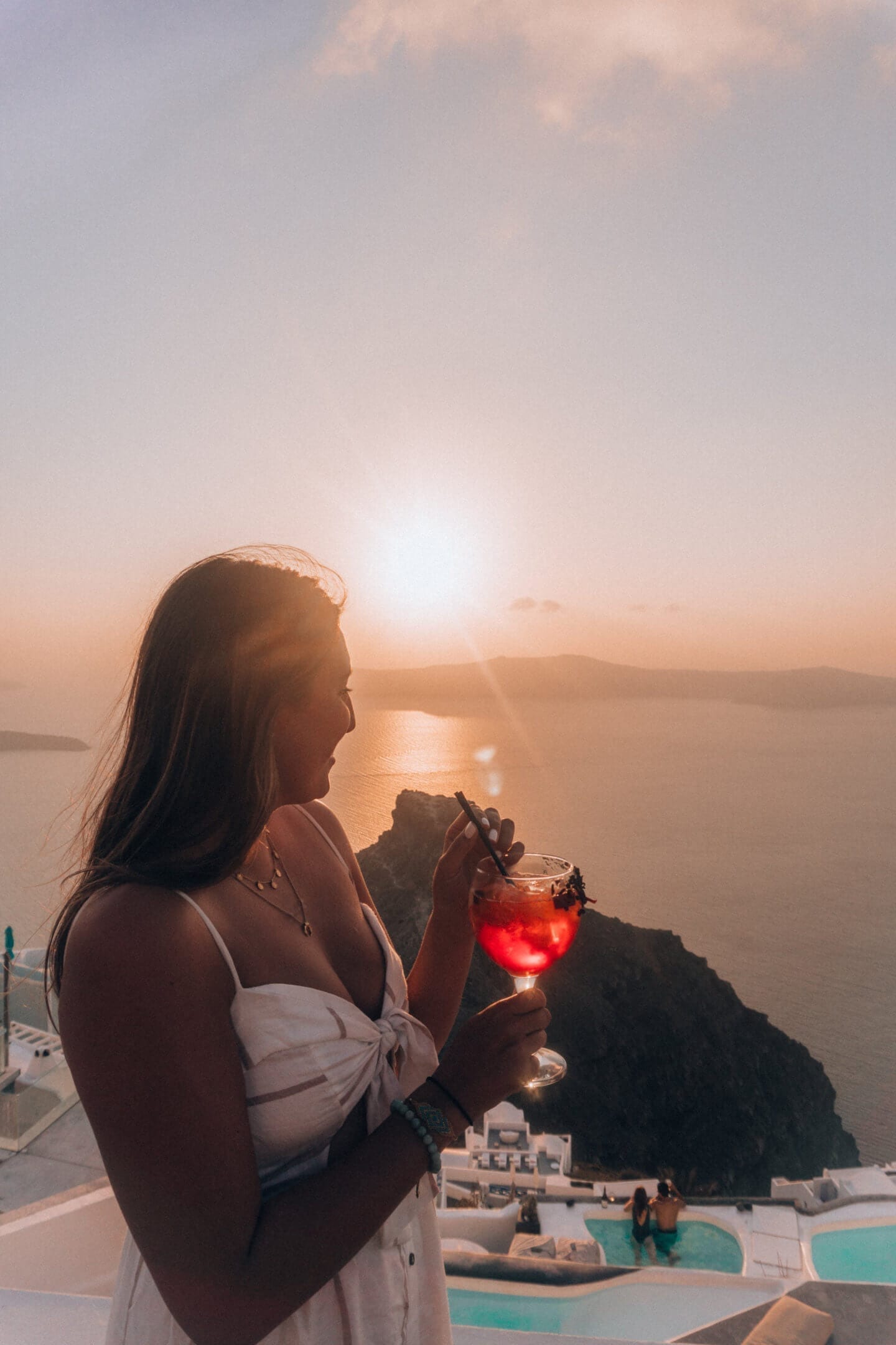 A girl looks out towards the sunset in Santorini holding an Aperol Spritz