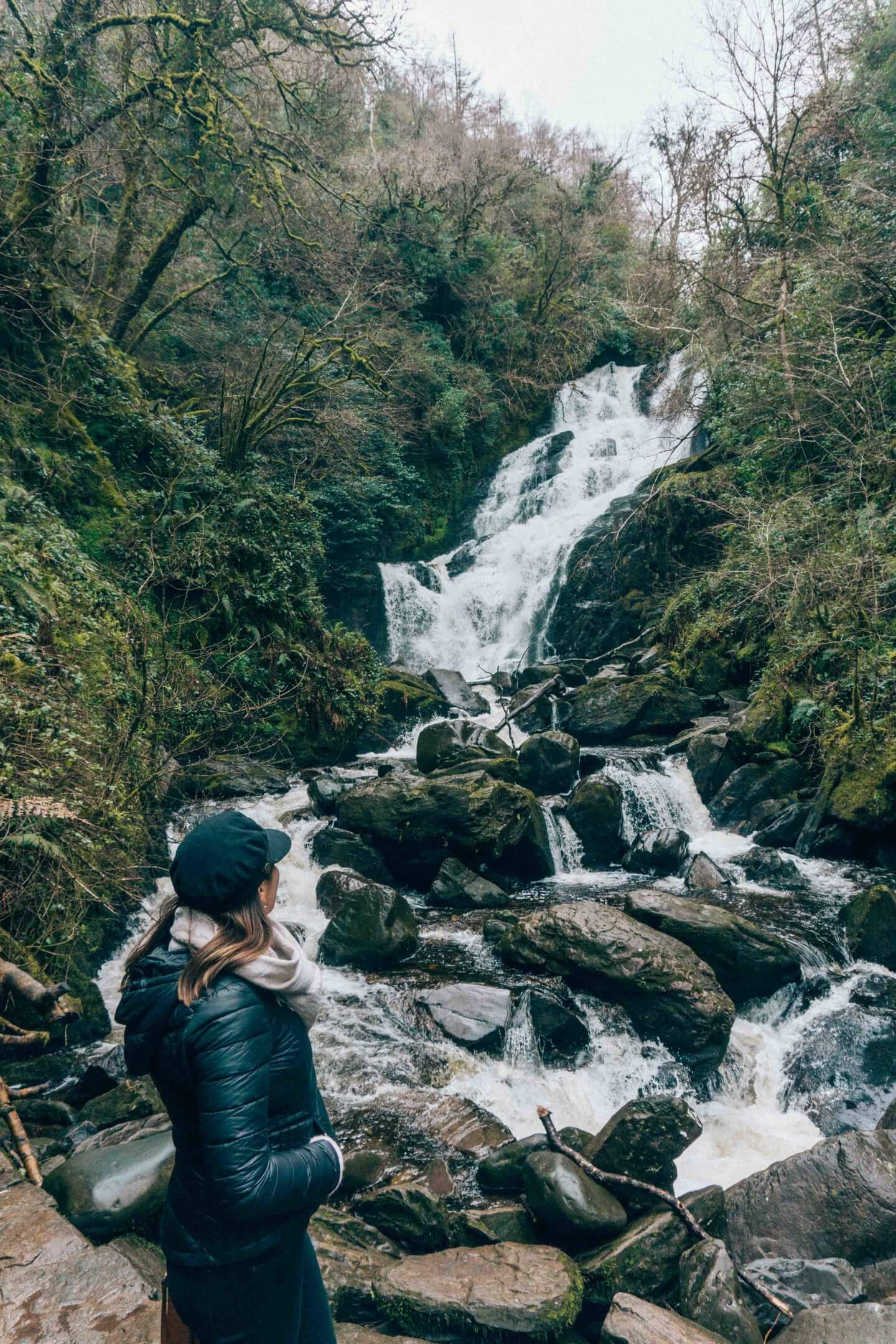 A girl with a hat looking up at the waterfall in Killarney National Park 