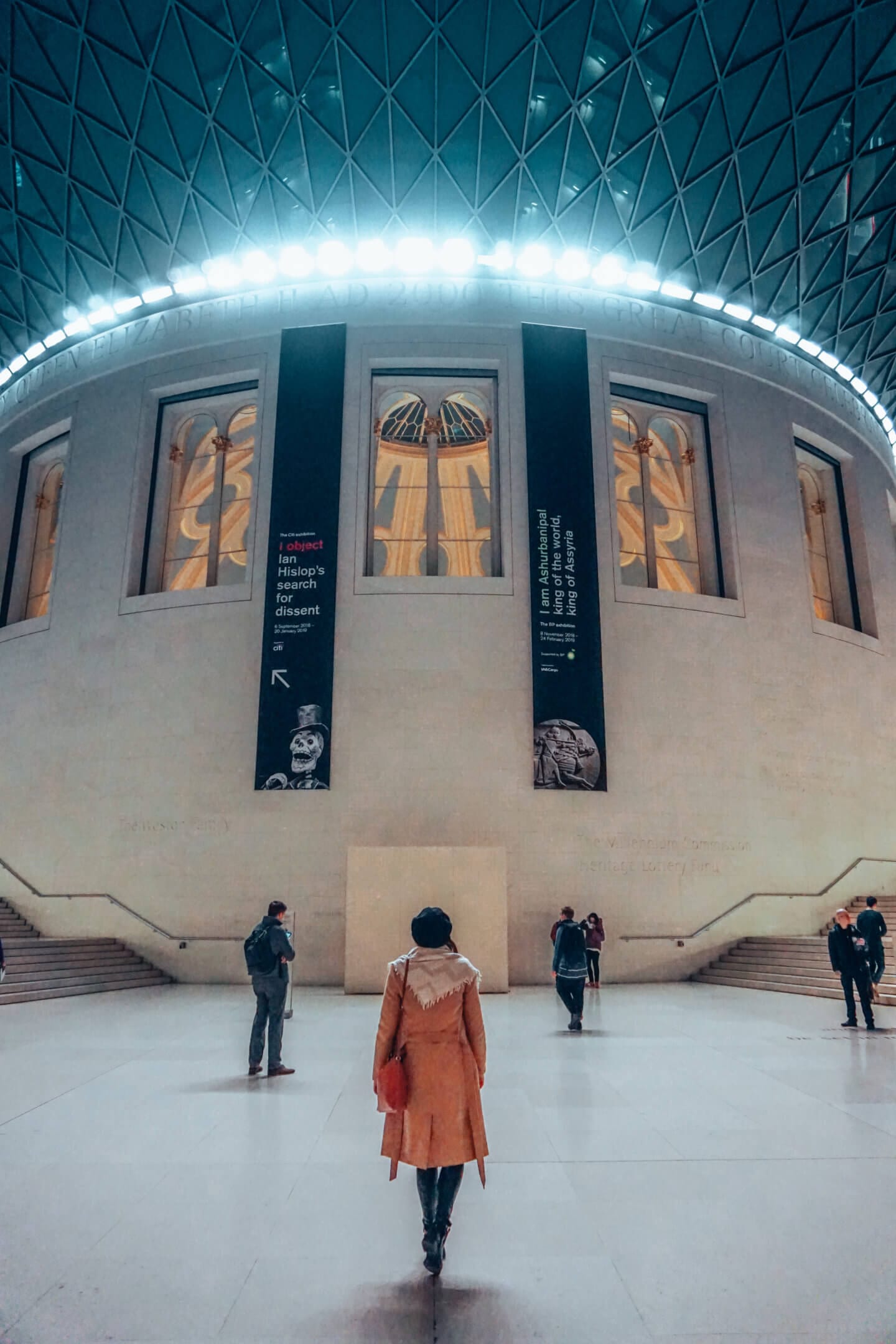 A girl wearing a black hat and long brown coat looks up at the British Museum in London