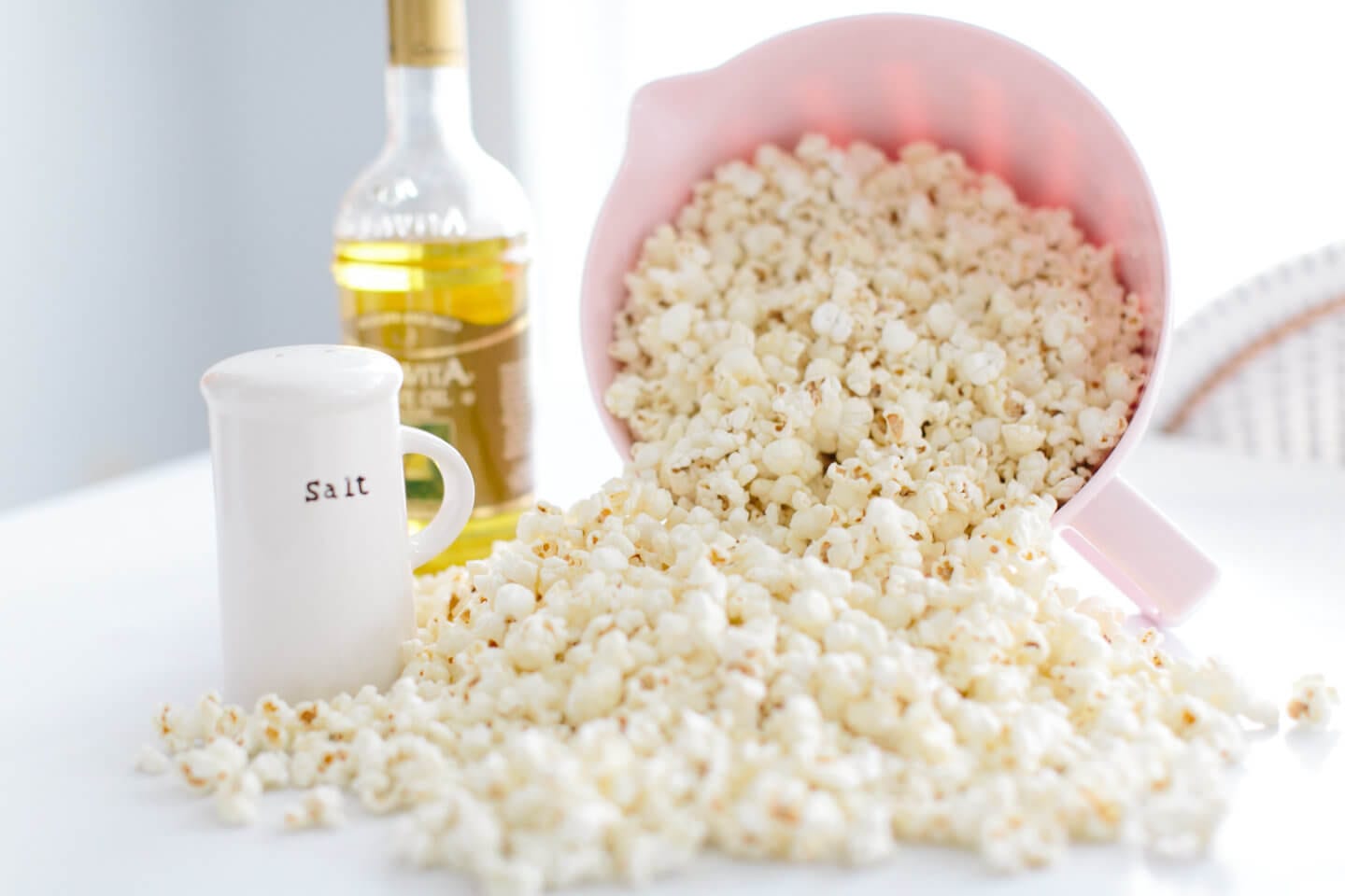 A pink bowl of popcorn spills out beside a bottle of olive oil and salt