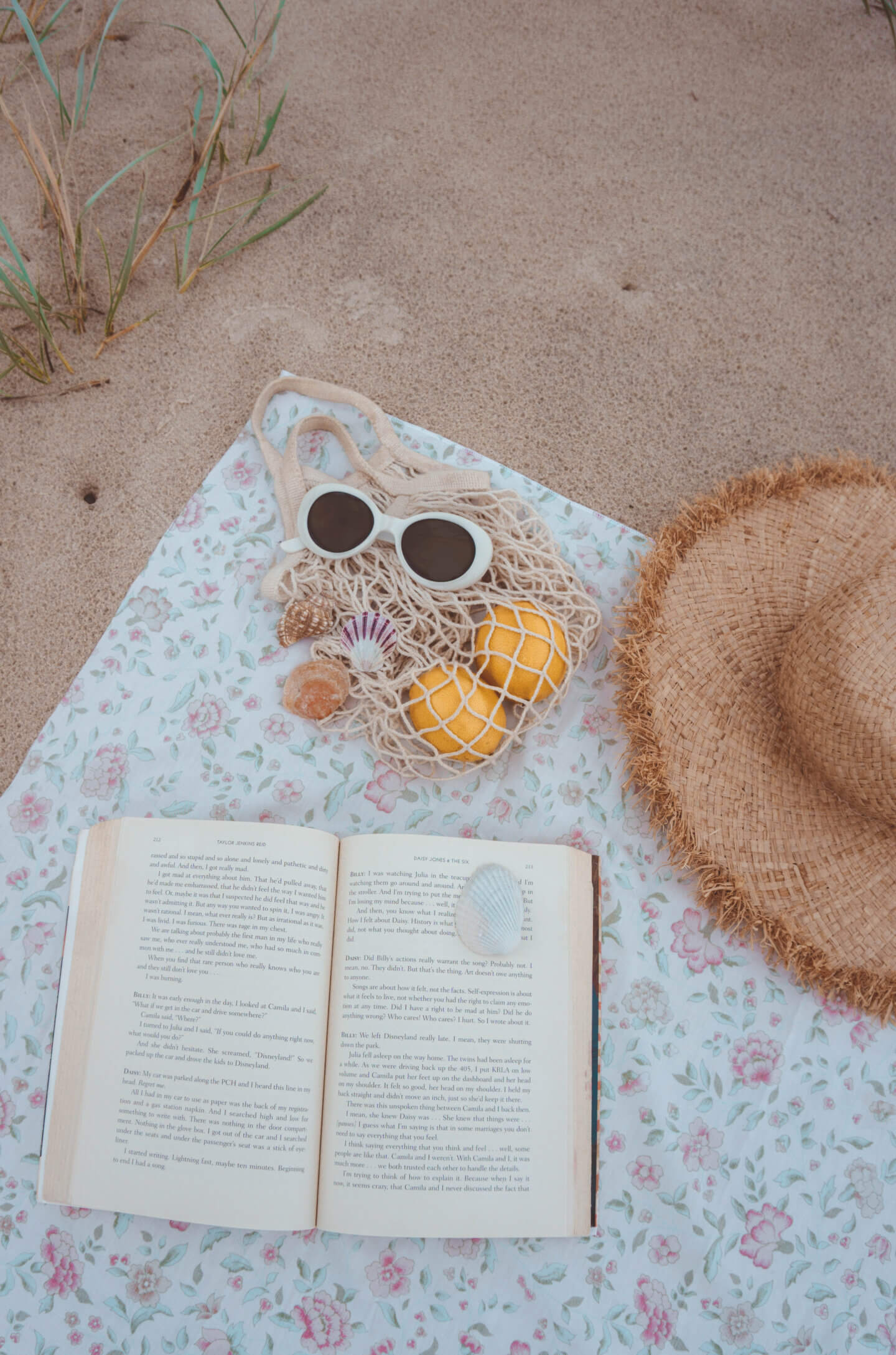 An open travel book on top of a floral beach towel with a hat, sunglasses, and shells