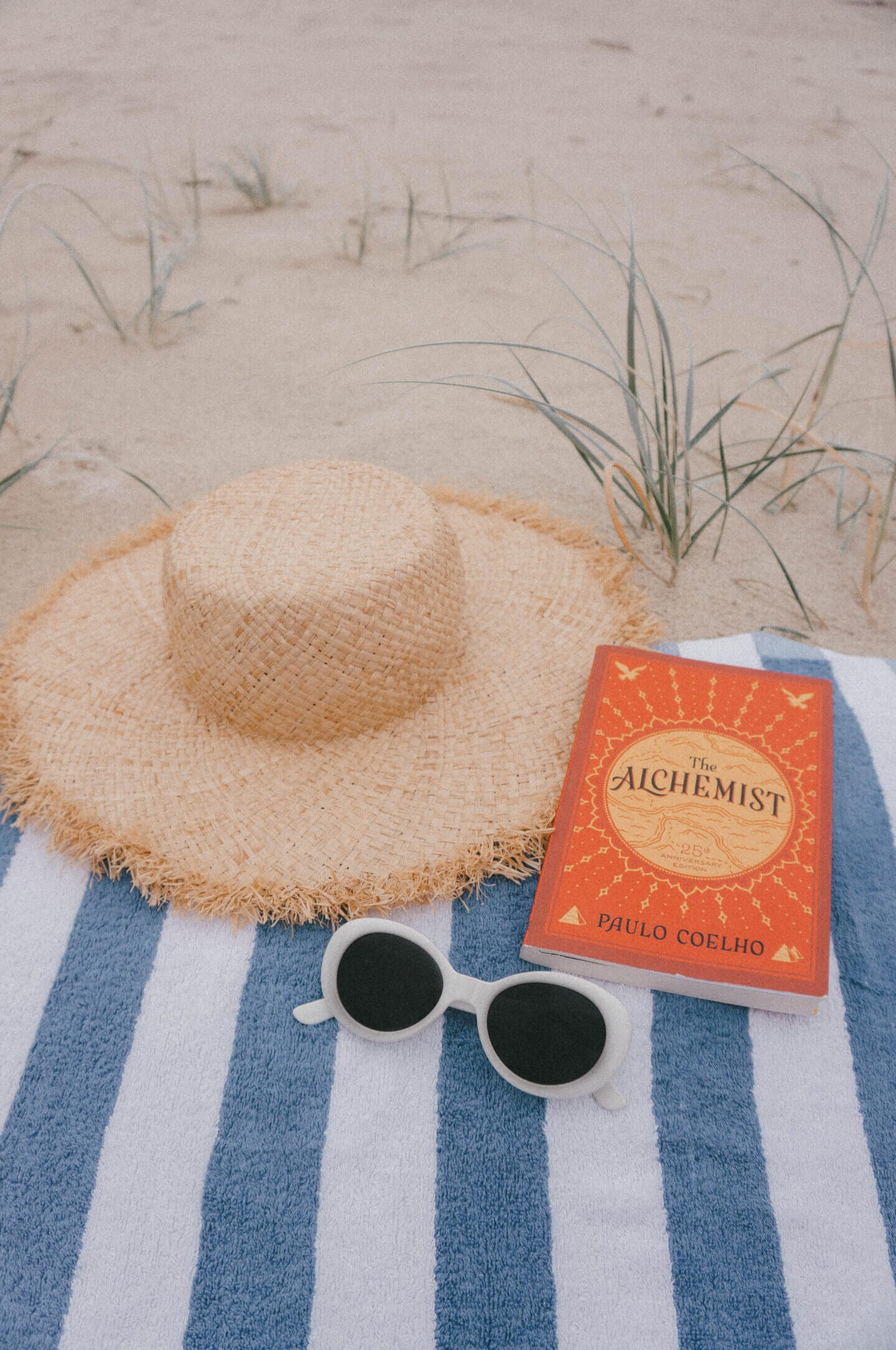 The Alchemist, a travel book on top of a beach towel with a hat and white sunglasses