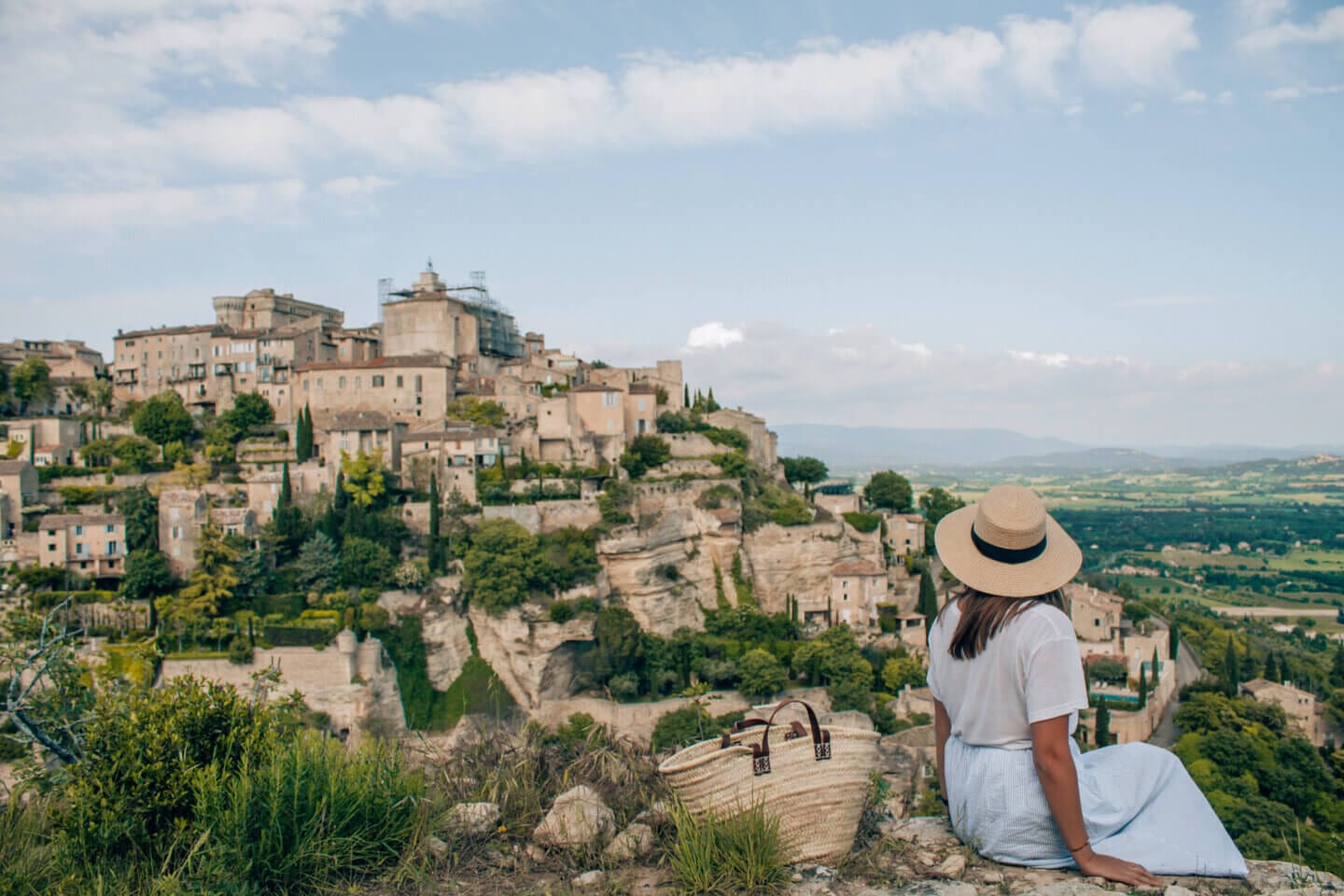 A girl with a hat on sits on a cliff and admires the beautiful small town of Gordes in Provence, France. One of the Most Romantic Places in Europe