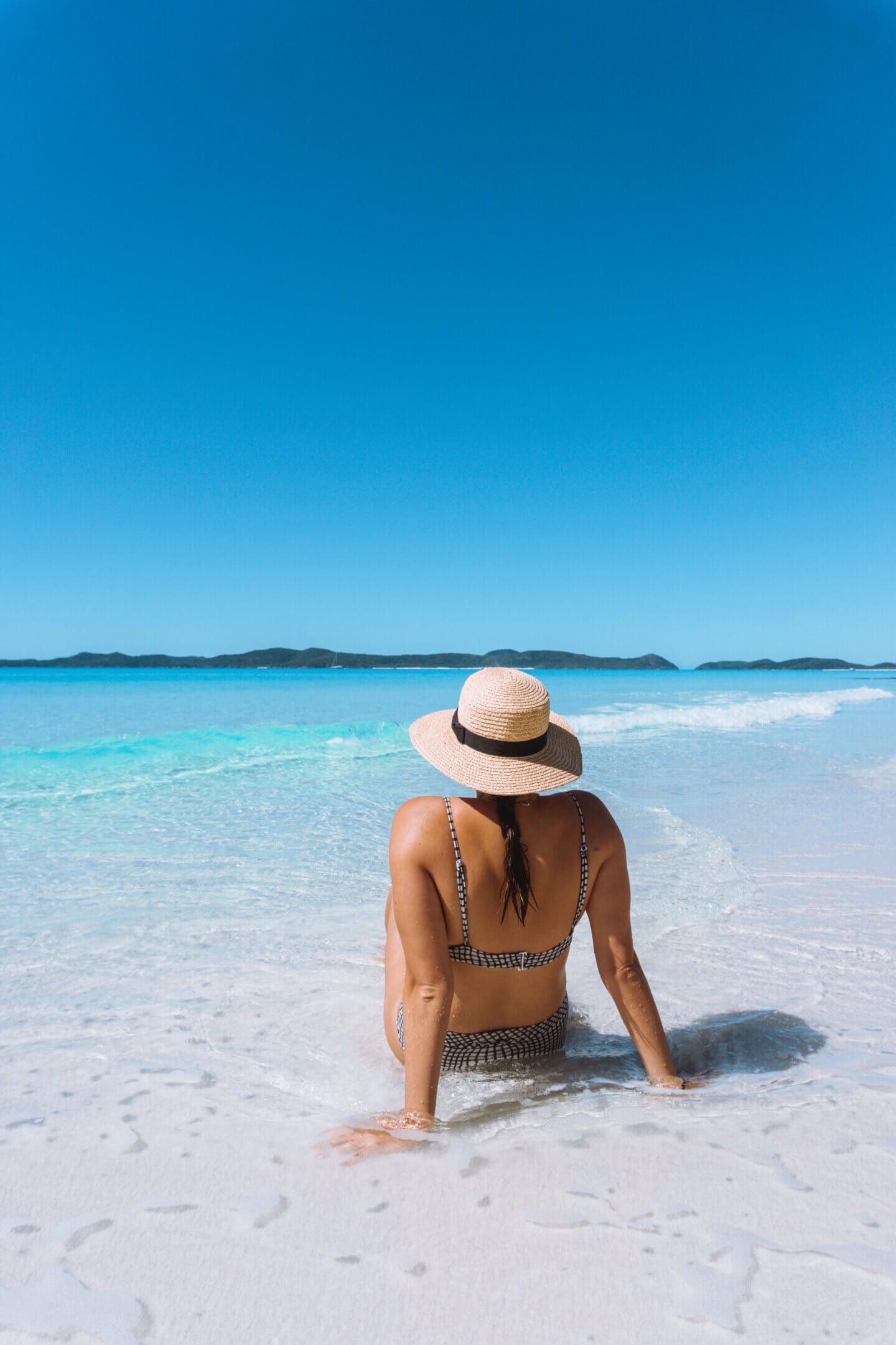 A girl with a hat on sits on Whitehaven Beach as the tide comes in