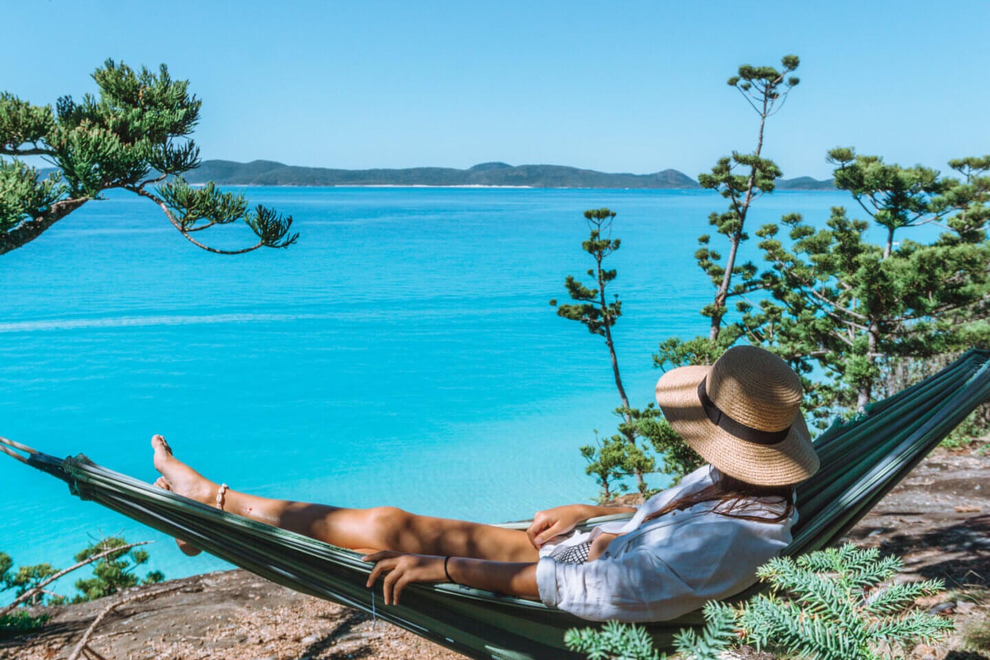 A girl with a hat on looks out towards Whitehaven Beach from a hammock