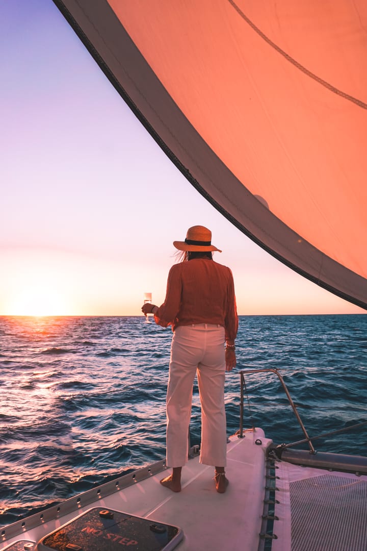 A girl with a hat on looks out towards sunset with a glass of champagne in hand.