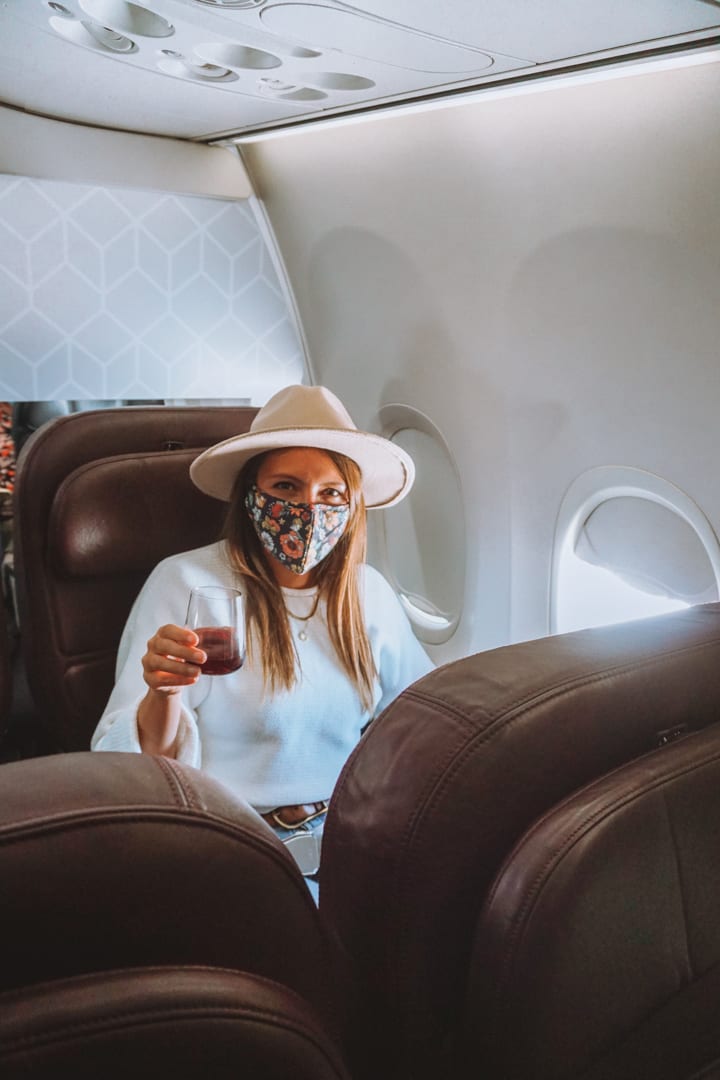 Girl wearing a hat and mask sits on a plane with a glass of red wine to Alice Springs for a road trip to Uluru