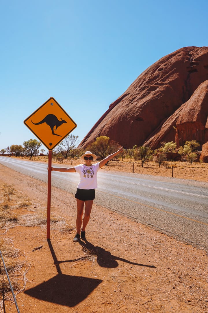 Alice Springs to Uluru Road Trip; Girl with a hat holds onto a kangaroo road sign beside Uluru
