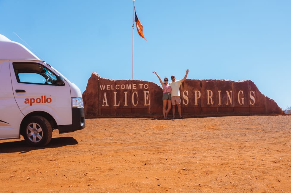Couple stand in front of the Welcome to Alice Springs sign on their way to Uluru with their Apollo campervan 
