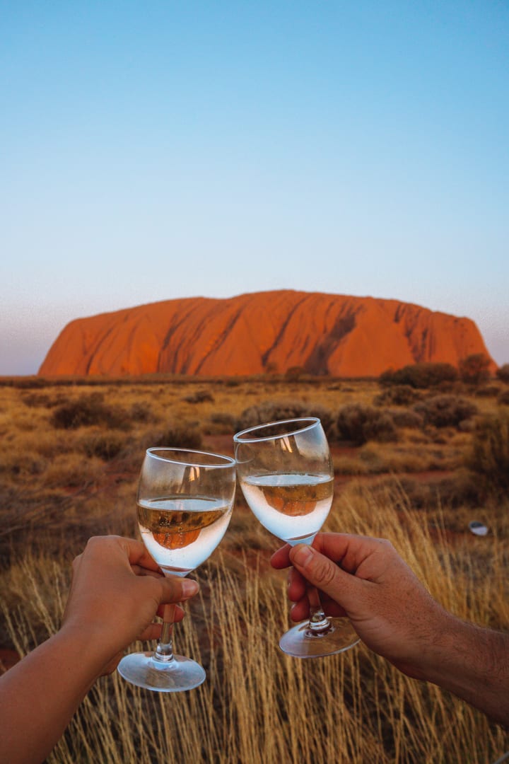 Alice Springs to Uluru Road trip: Two people cheersing champagne in front of Uluru at sunset