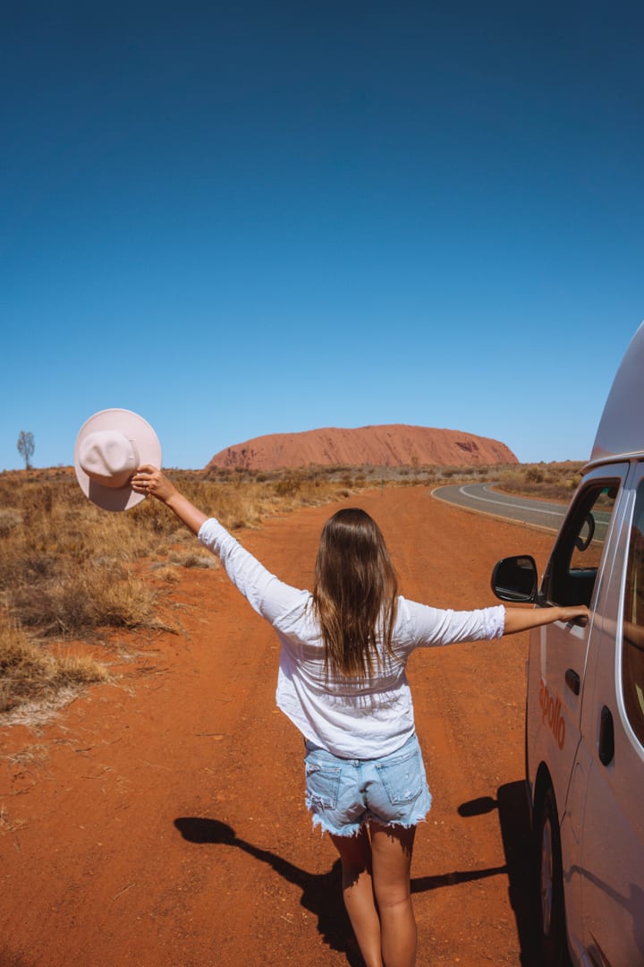 Alice Springs to Uluru Road Trip: Girl in front of the road to Uluru holding onto a campervan and a hat.