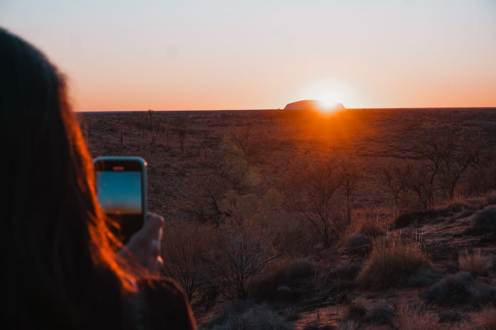 Girl talks a beautiful photo with her smart phone of the golden sun rising above Uluru.