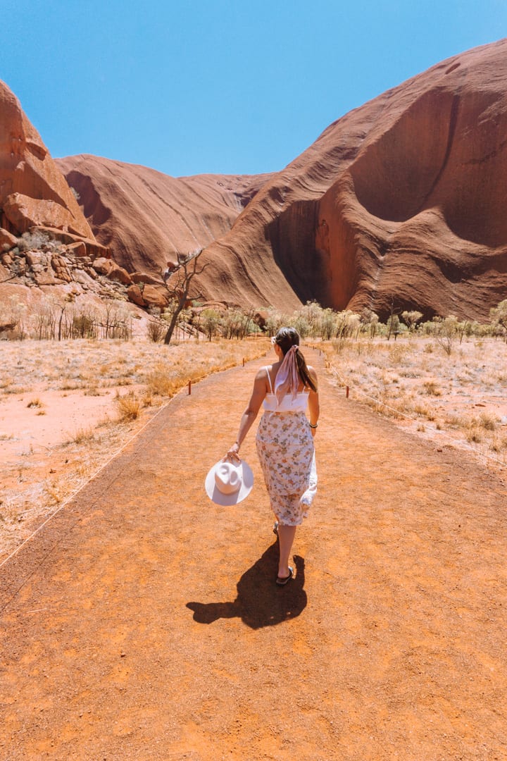 Girl walks towards Uluru with a hat in hand.