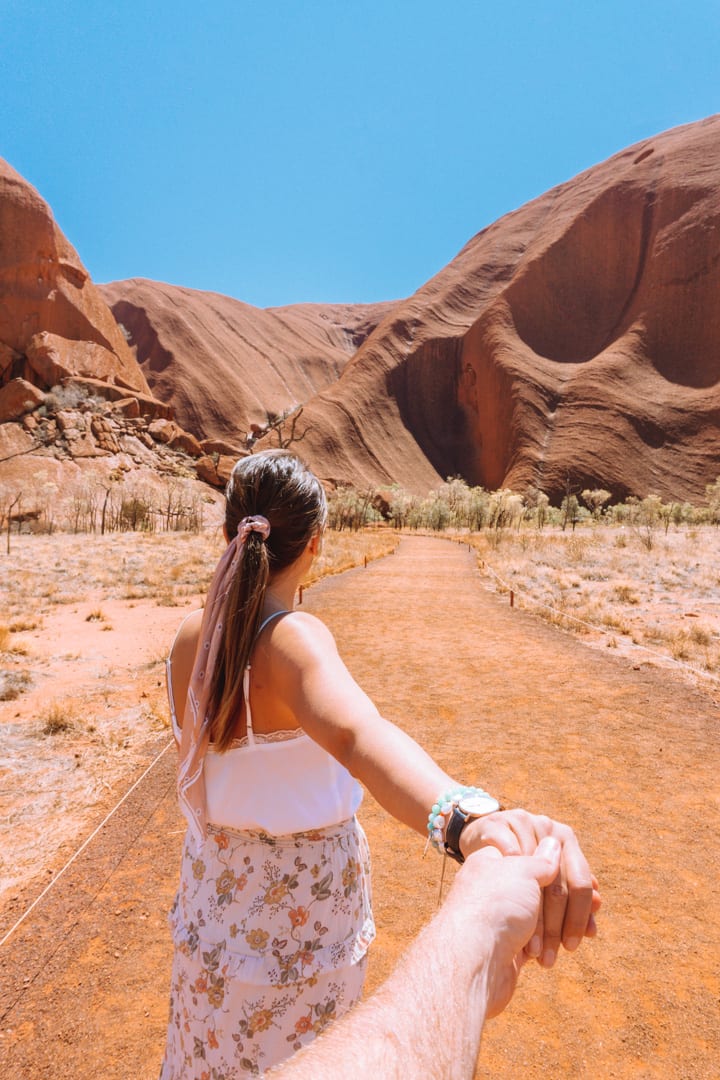 A girl with a ribbon in her hair holds the hand of her partner while looking at Uluru