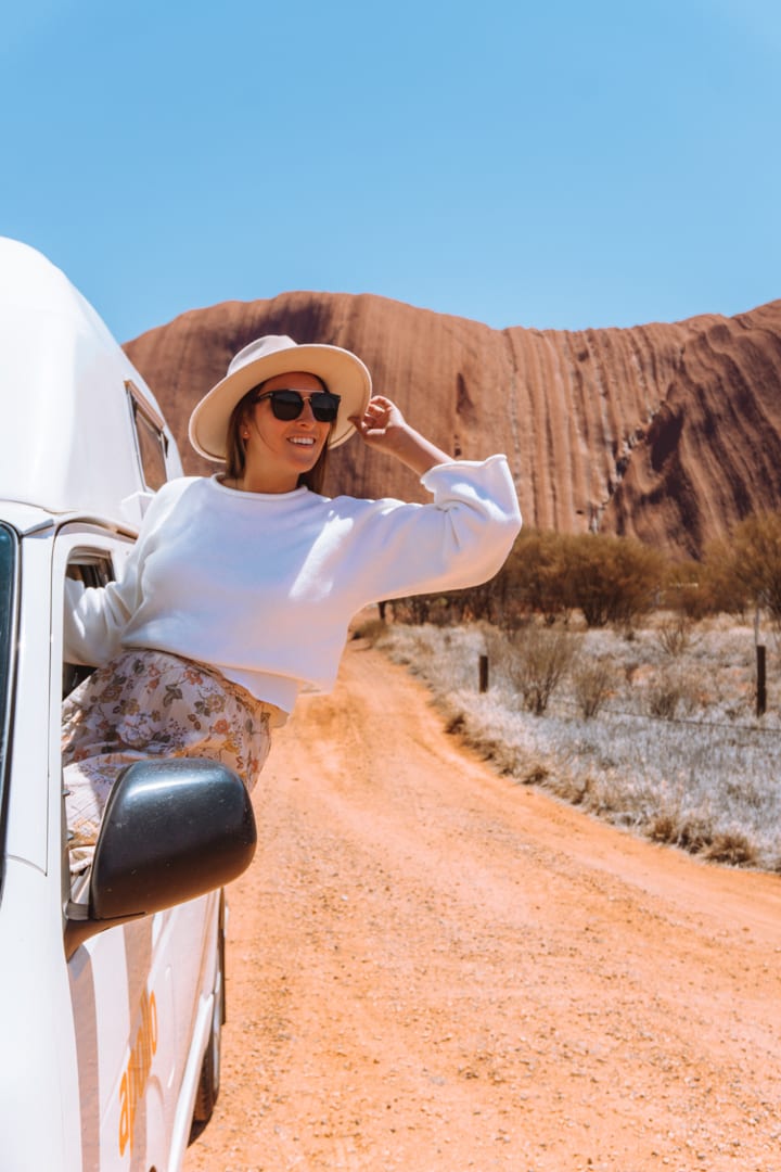 Alice Springs to Uluru Road Trip: A girl holds onto her hat while sitting out the window of her campervan in front of Uluru