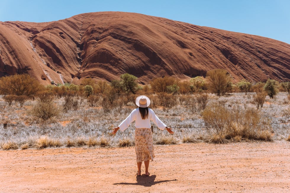 Alice Springs to Uluru Road Trip: Girl with a hat on looking up at Uluru with her arms out