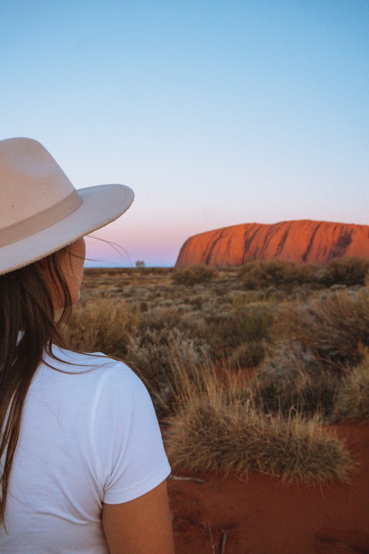Girl with a hat on looks out at Uluru at sunset at the dune walk
