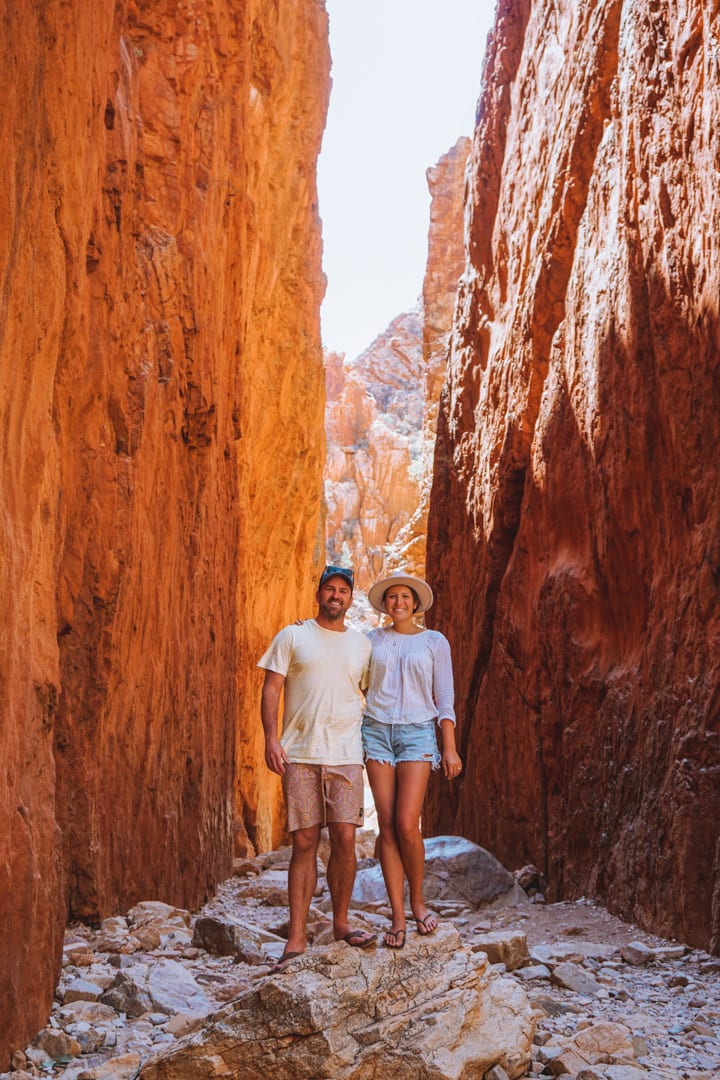 Alice Springs to Uluru Road Trip: A couple, both wearing hats, standing in Standley Chasm at noon, when the walls are glowing bright orange.