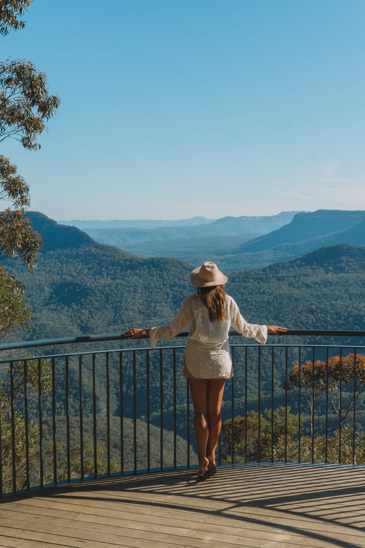 A girl wearing a hat and white shirt look out over the Blue Mountain range and at the Megalong Valley below. 