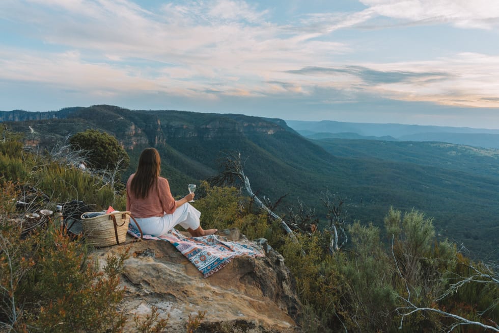 Best places to Eat in the Blue Mountains: A girl sits on a blanket with a picnic basket and glass of wine while looking out at the Blue Mountains, Australia