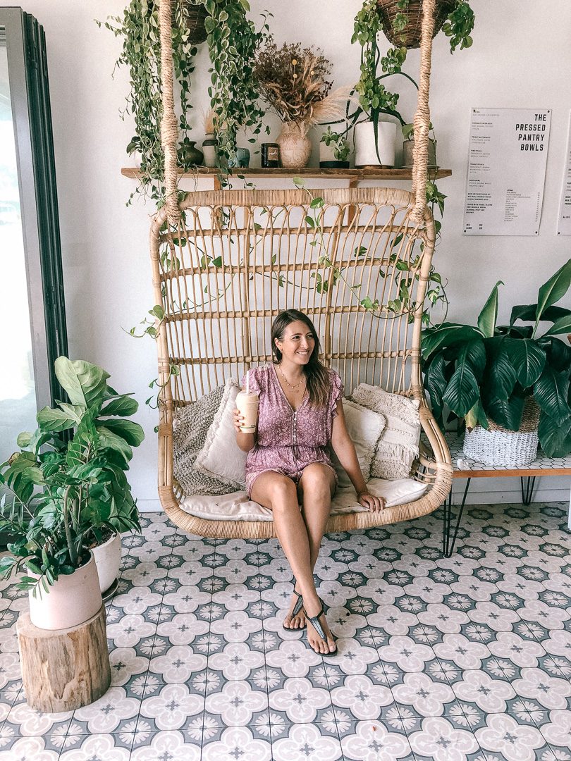 A girl sitting on a rattan swinging chair surrounded by plants in the cafe Pressed Pantry in Byron Bay