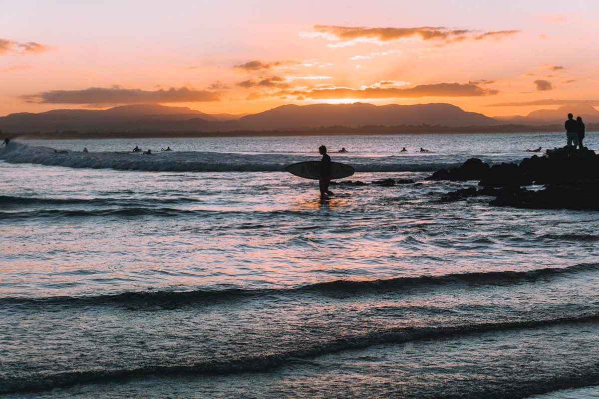 A bright pink and orange sunsest lights up the sky as a surfer walks into the water at the Pass in Byron Bay, Australia.