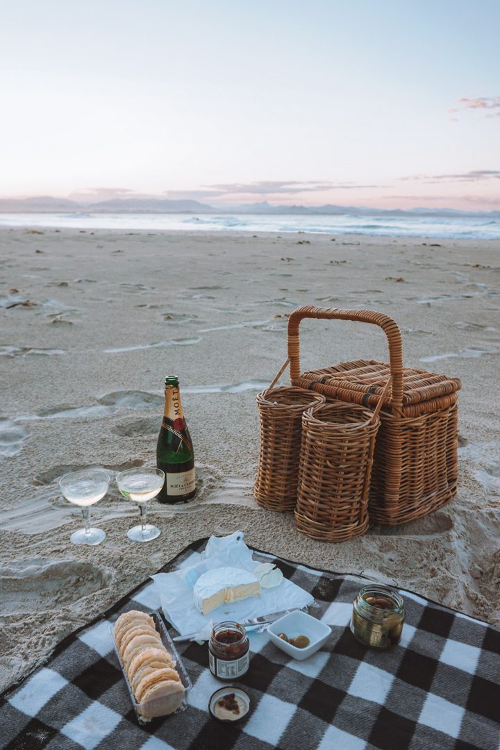 A picnic basket, blanket, snacks and champagne sit out on the sand in Byron Bay, Australia.