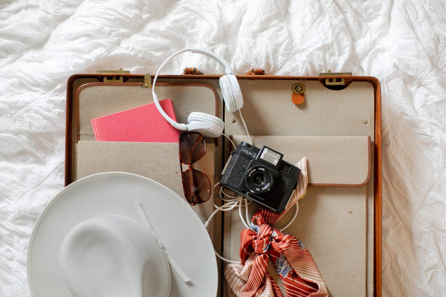 Looking down at a ladies suitcase with a hat, scarf, sunglasses, camera, journal and headphones. Best Travel Planning Resources & Travel Gear I Swear By