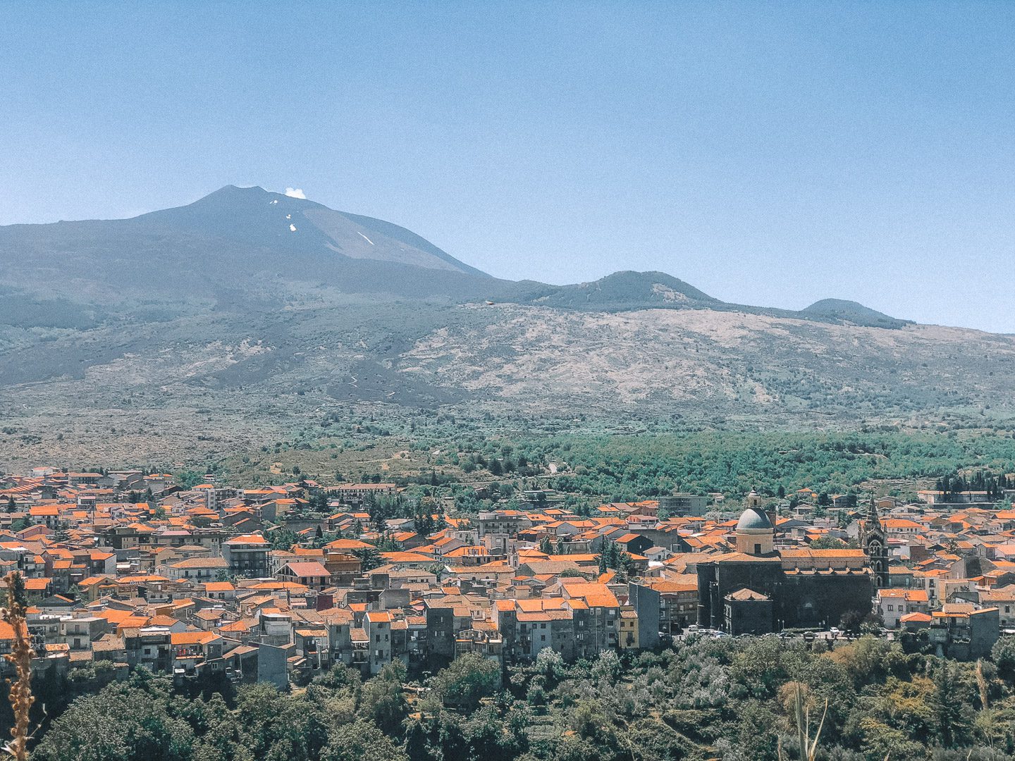 Mount Etna lets out a puff of smoke on a clear blue bird day, with the small town of Catania at it's feet - a must do on this 7-day Sicily Itinerary