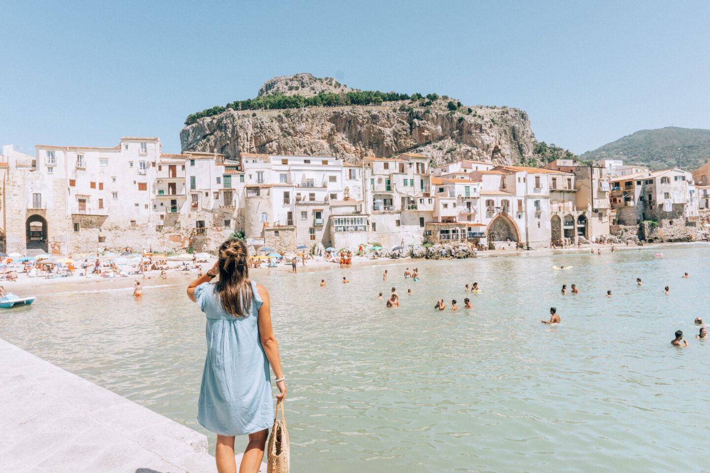 A girl with a blue dress on looks back towards the beach, La Rocca mountain and the seaside town of Cefalù in Sicily