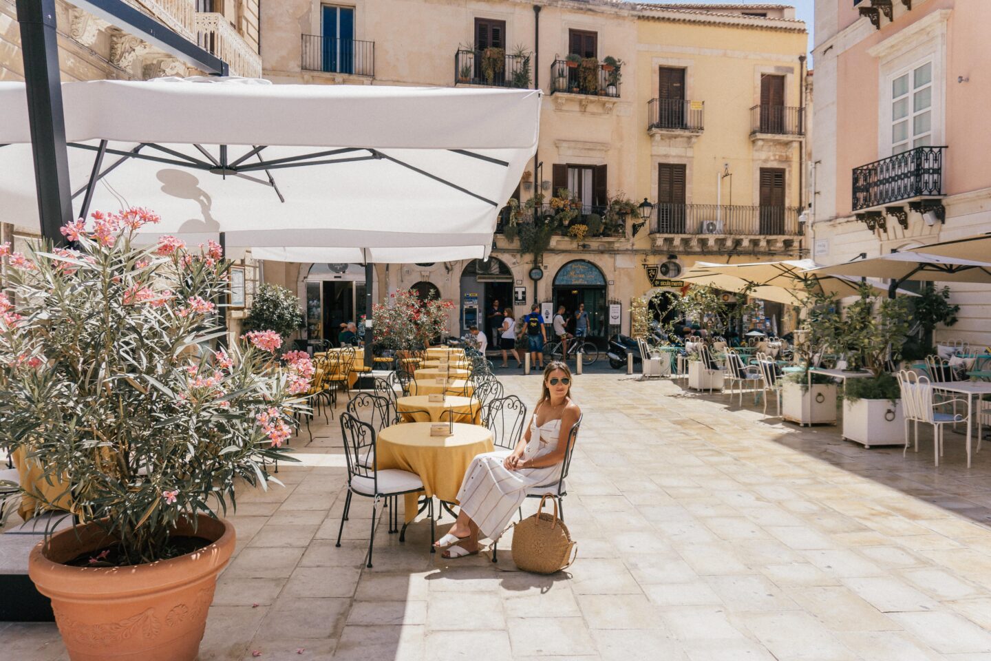 A girl wearing a white dress sits at a European cafe during the day, admiring the beautiful town of Oritgia - a hidden gem on this 7-day Sicily Itinerary
