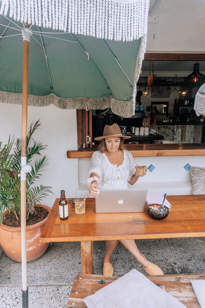 A girl in a white dress and hat sips on a coffee and looks at her computer on the patio at the Byron Bay General Store