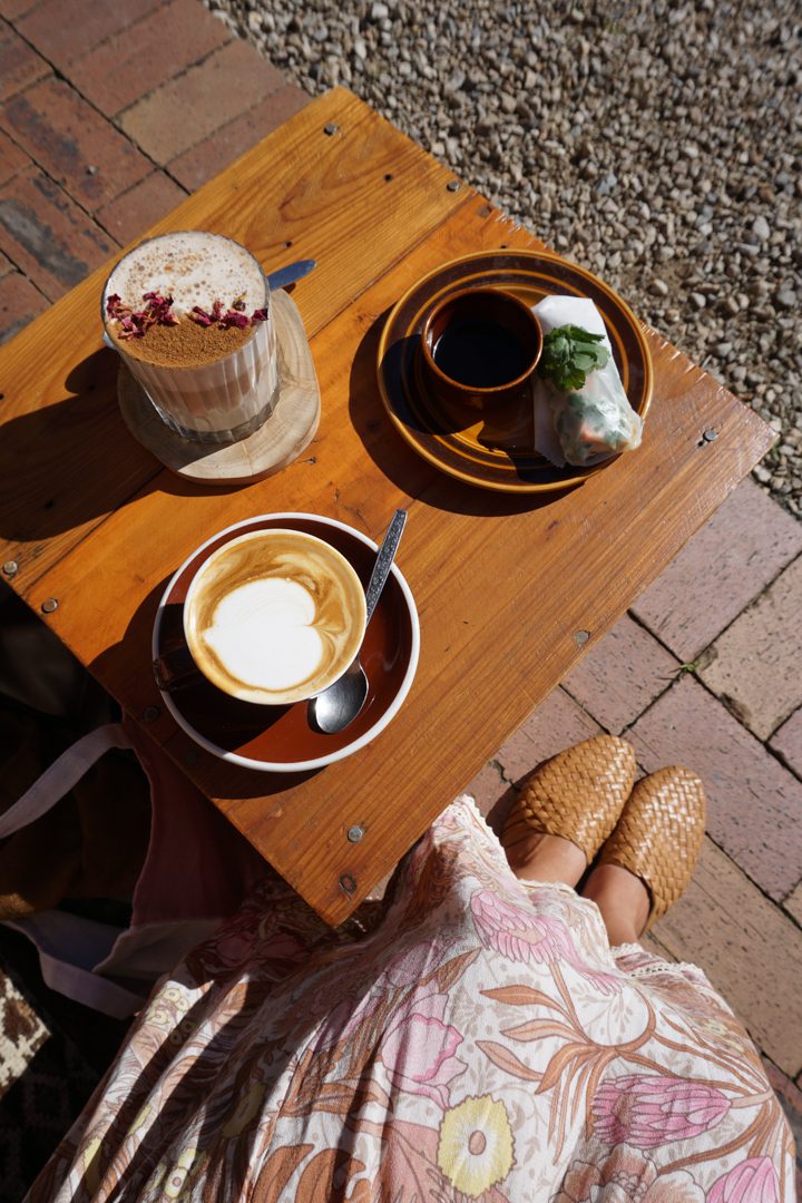 A girl wearing a dress looks down at a flat white, tea and snack on a table at Folk cafe in Byron Bay