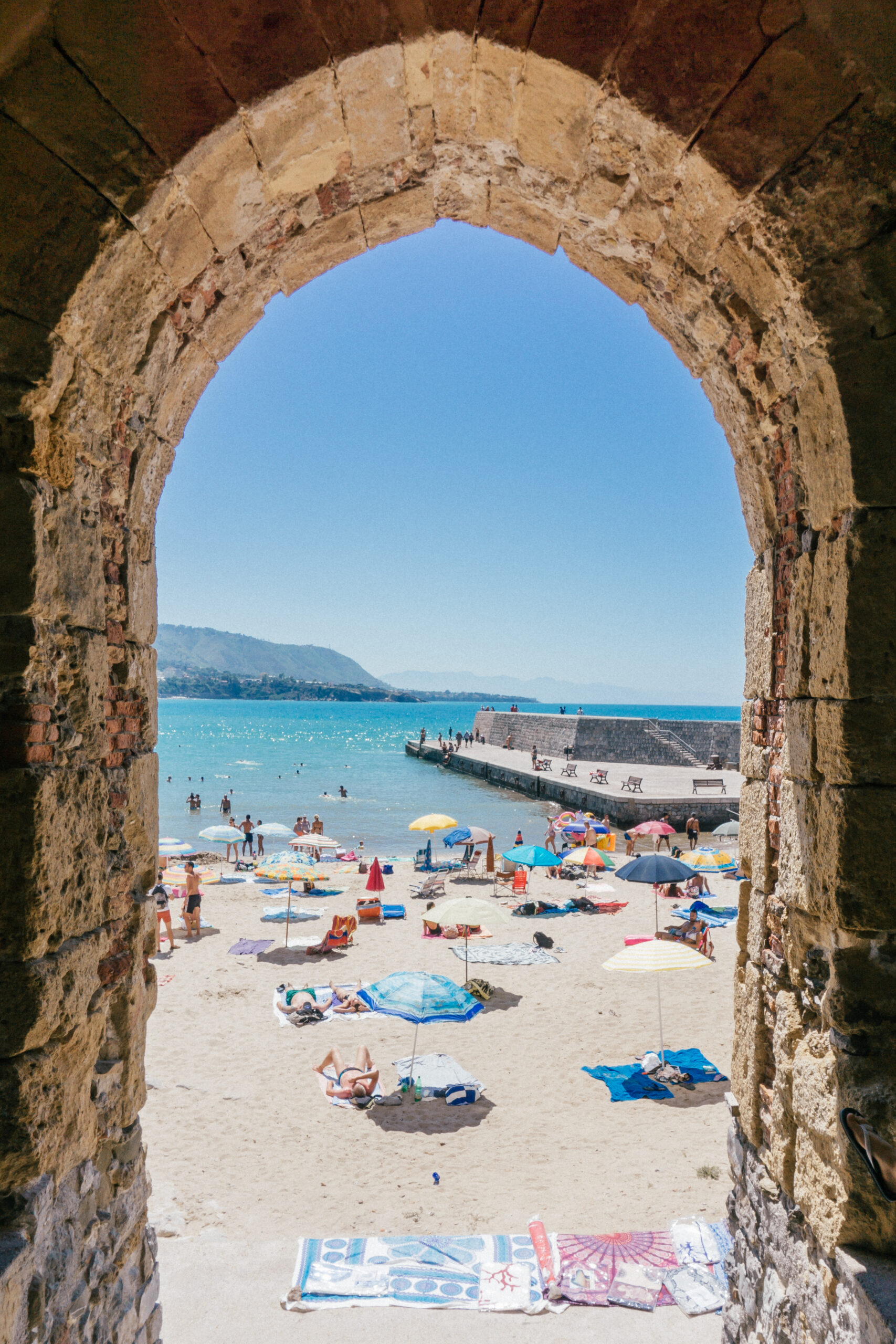 Looking through an brick archway to the beach and colourful umbrellas in  Cefalù - a gorgeous stop on this 7-day Sicily Itinerary