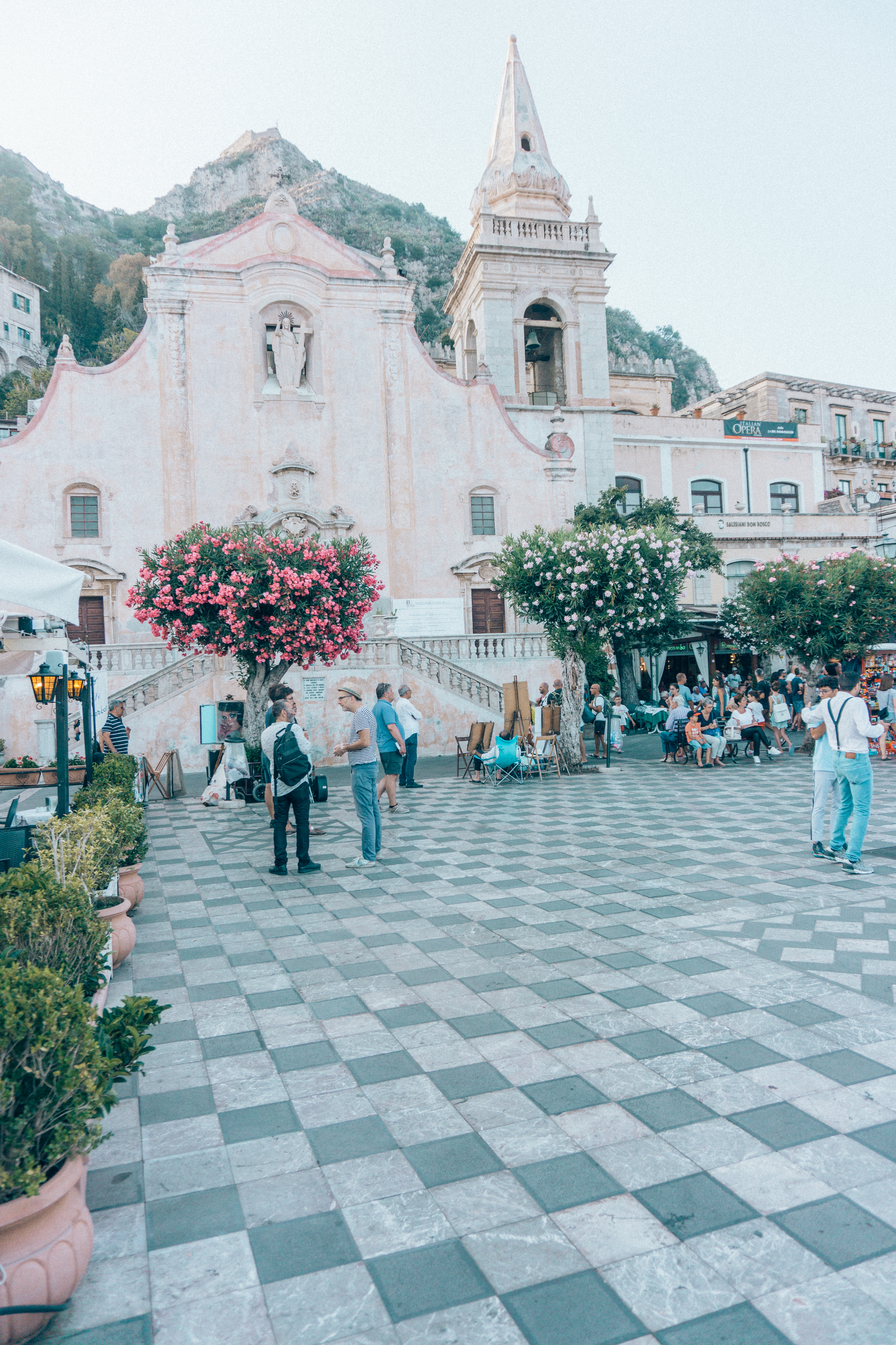 People hanging out at the cliff top square of Taormina with the pink cathedral and bell tower, along with the checkered pavement. 