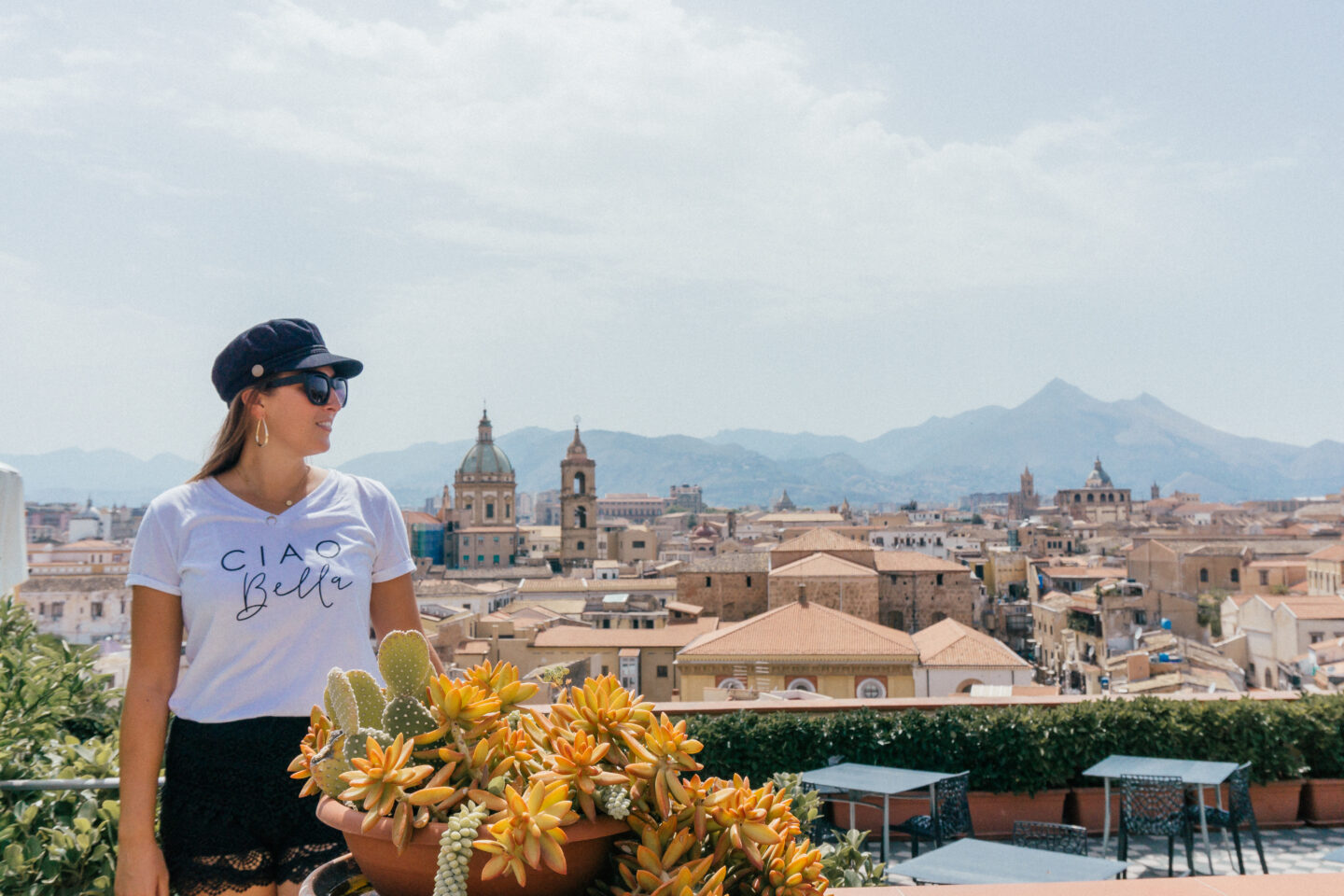 A girl wearing a hat sits on the Seven Restaurant rooftop patio admiring the ancient city of Palermo in Sicily during the day.