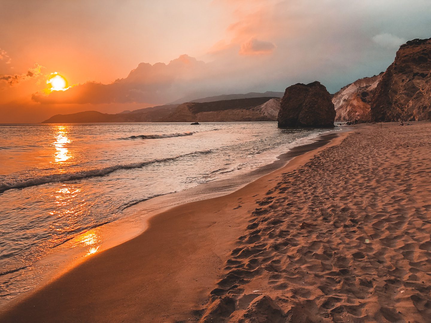 A iconic orange sunset reflects on he sandy shores of Firiplaka Beach in Milos Island, Greece
