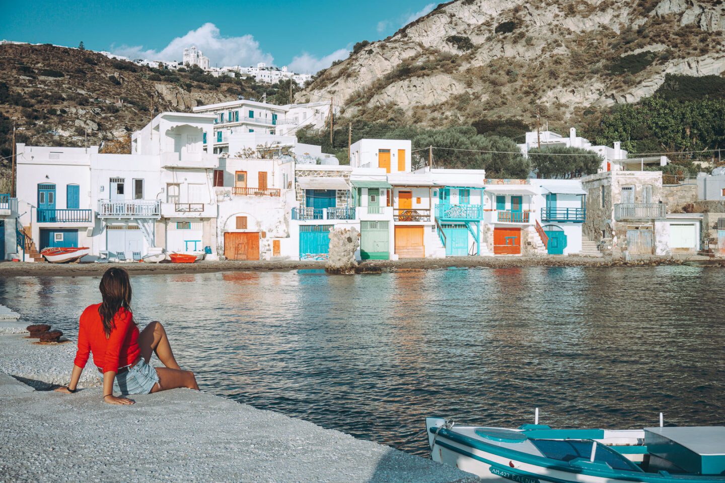 A girl in a red sweater sits on the jetty and admires the colourful fishing village of Klima on Milos, when Greek Island Hopping