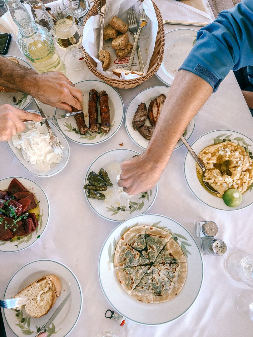 A table full of delicious Greek food and two people reaching to grab it while Greek Island Hopping