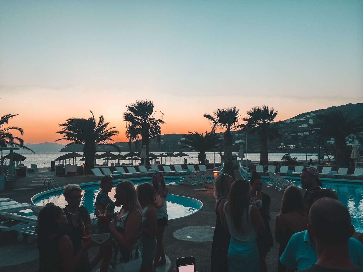 A group of people sip on cocktails while the sun sets at a beach bar on Ios Island in Greece