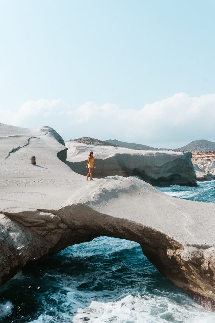 A girl in a yellow romper walks over an oceanside archway of Sarakiniko Beach on Milos Island in Greece. 