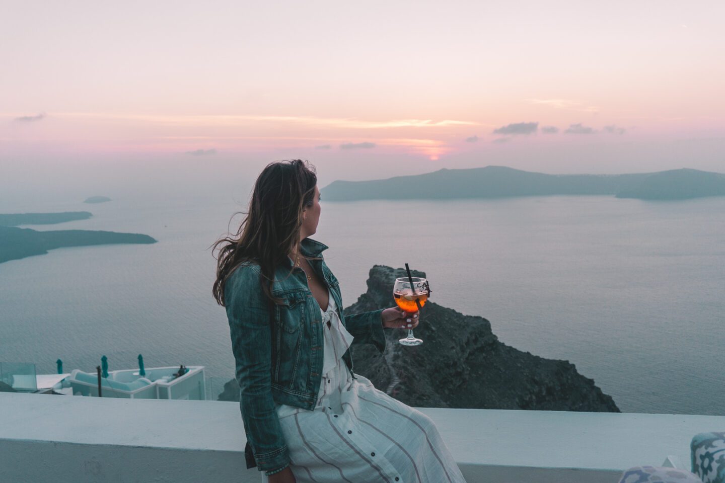 A girl holds a Aperol Spritz while watching the iconic Santorini sun set over the Greek Islands in the Aegean sea 