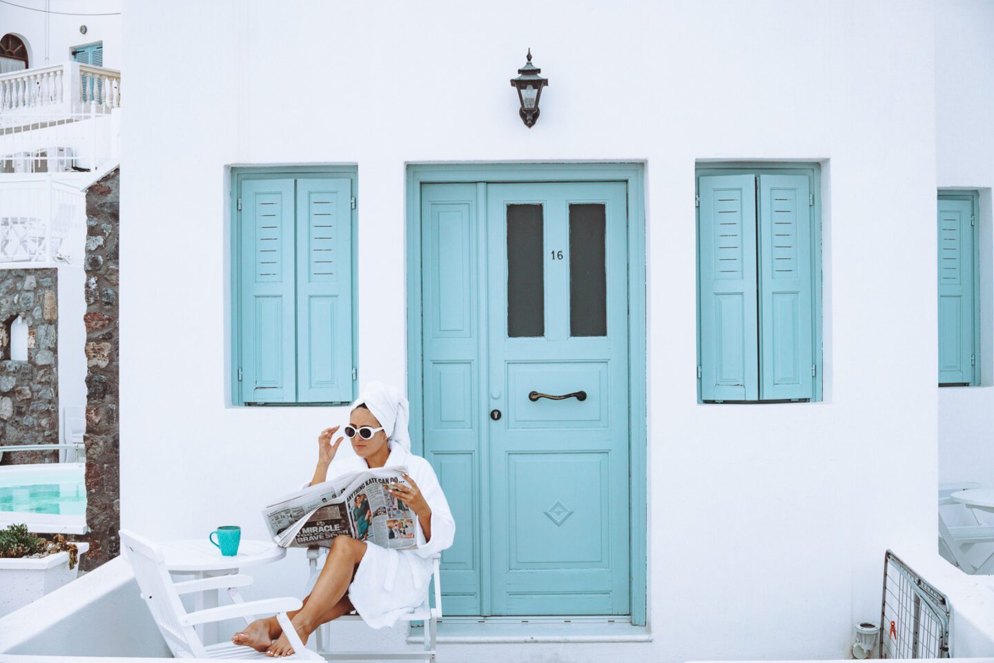 A girl reads the newspaper and enjoys a cup of coffee on her Greek patio in Santorini wearing a white bathrobe, sunglasses and a towel in her hair
