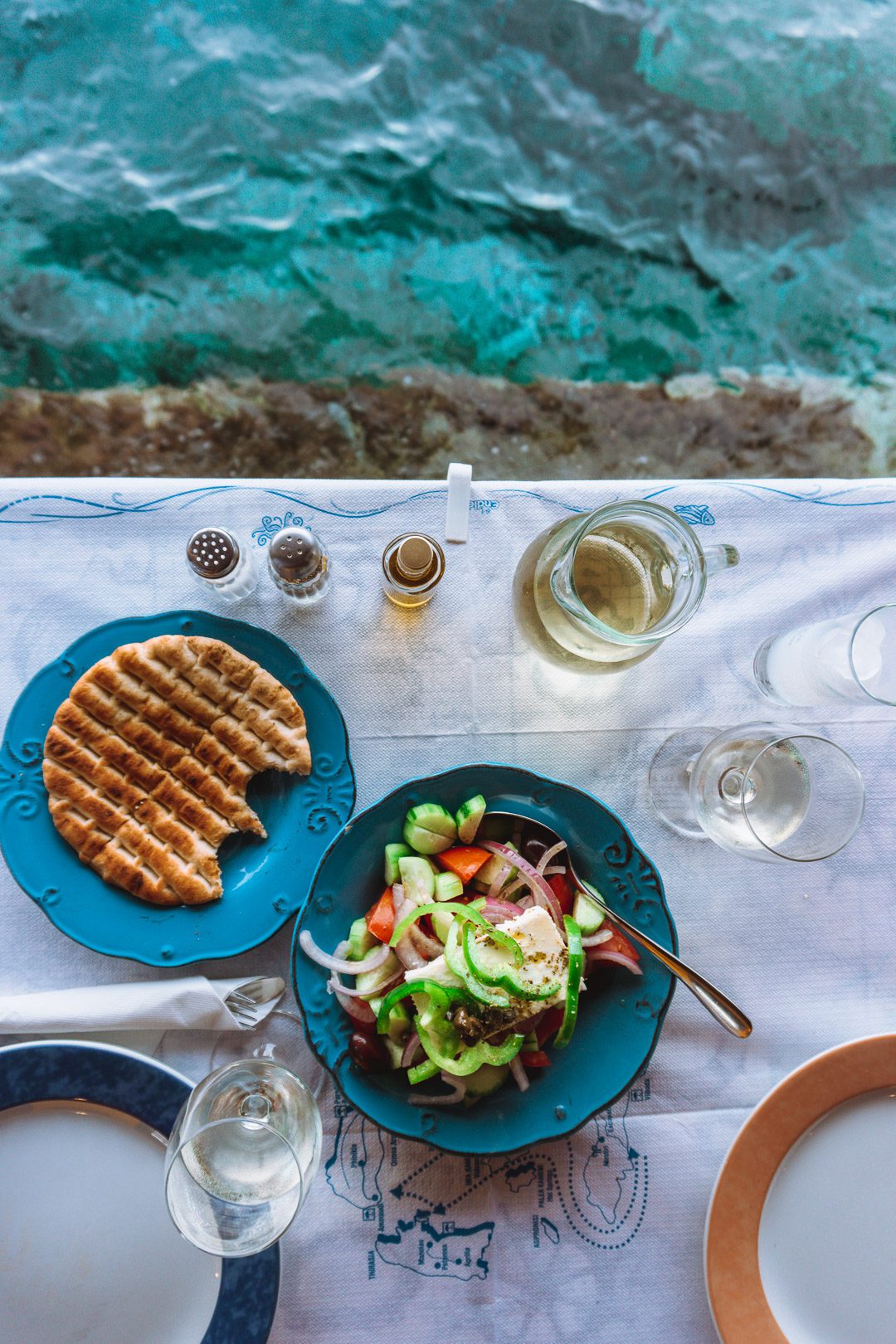 Looking down at an oceanside table with wine, pita bread, and a greek salad at the Seafood Tavern in Ammoundi Bay on Santorini Island