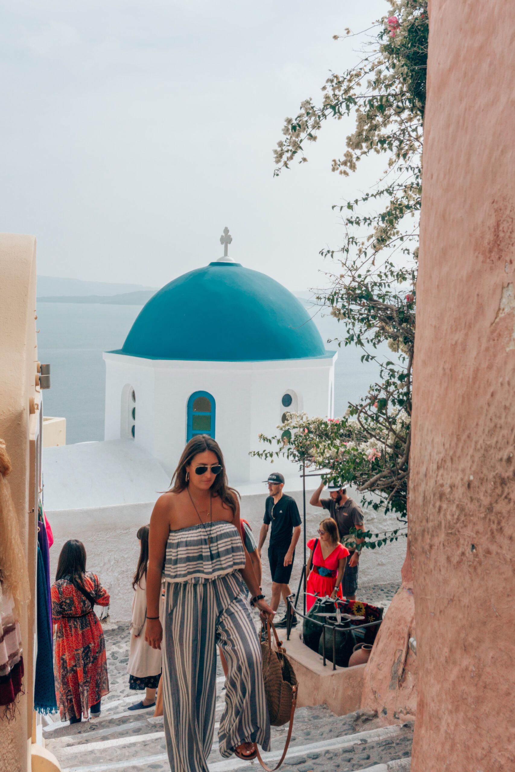 A girl wearing a stripped jumpsuit walks through the streets of Santorini, Greece with a classic blue domed church behind her. 