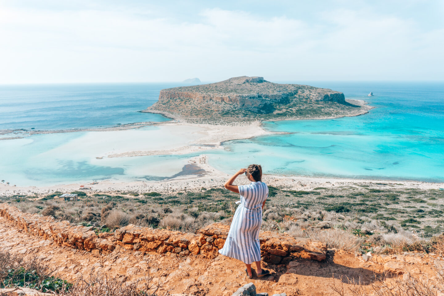 A girl on top of a rocky cliff looks out towards Balos Beach below on Crete, Greece. 