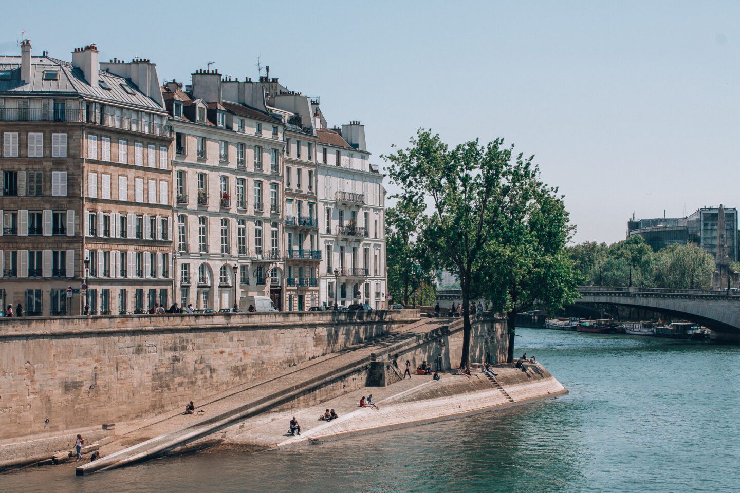 People sit along the Seine river in Paris, France soaking up the summer sunshine with classic Parisian apartment architecture in the background.