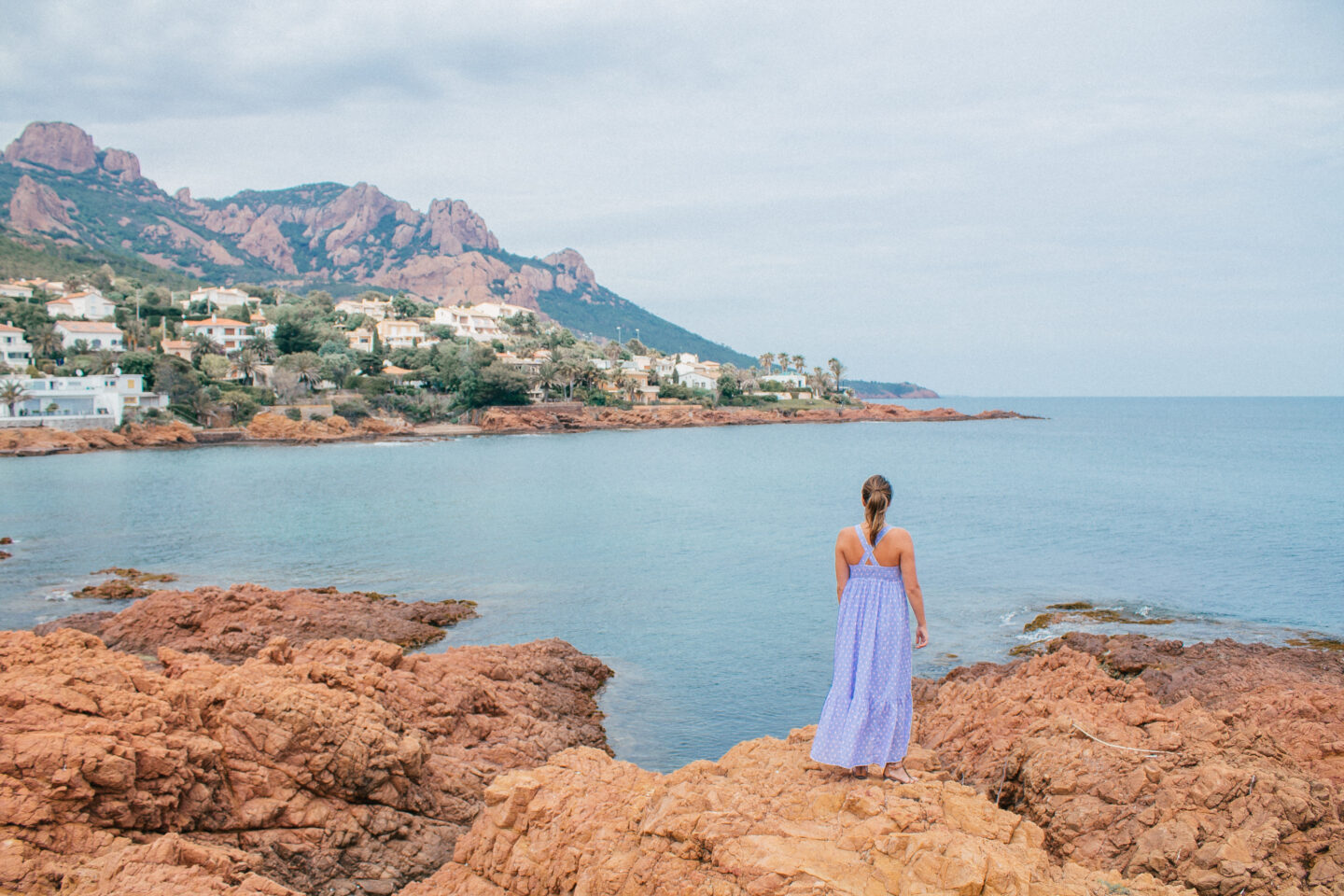A lady in a long blue dress walks along the red seaside rocks on the French Riveria, a must-see place to visit in France. 
